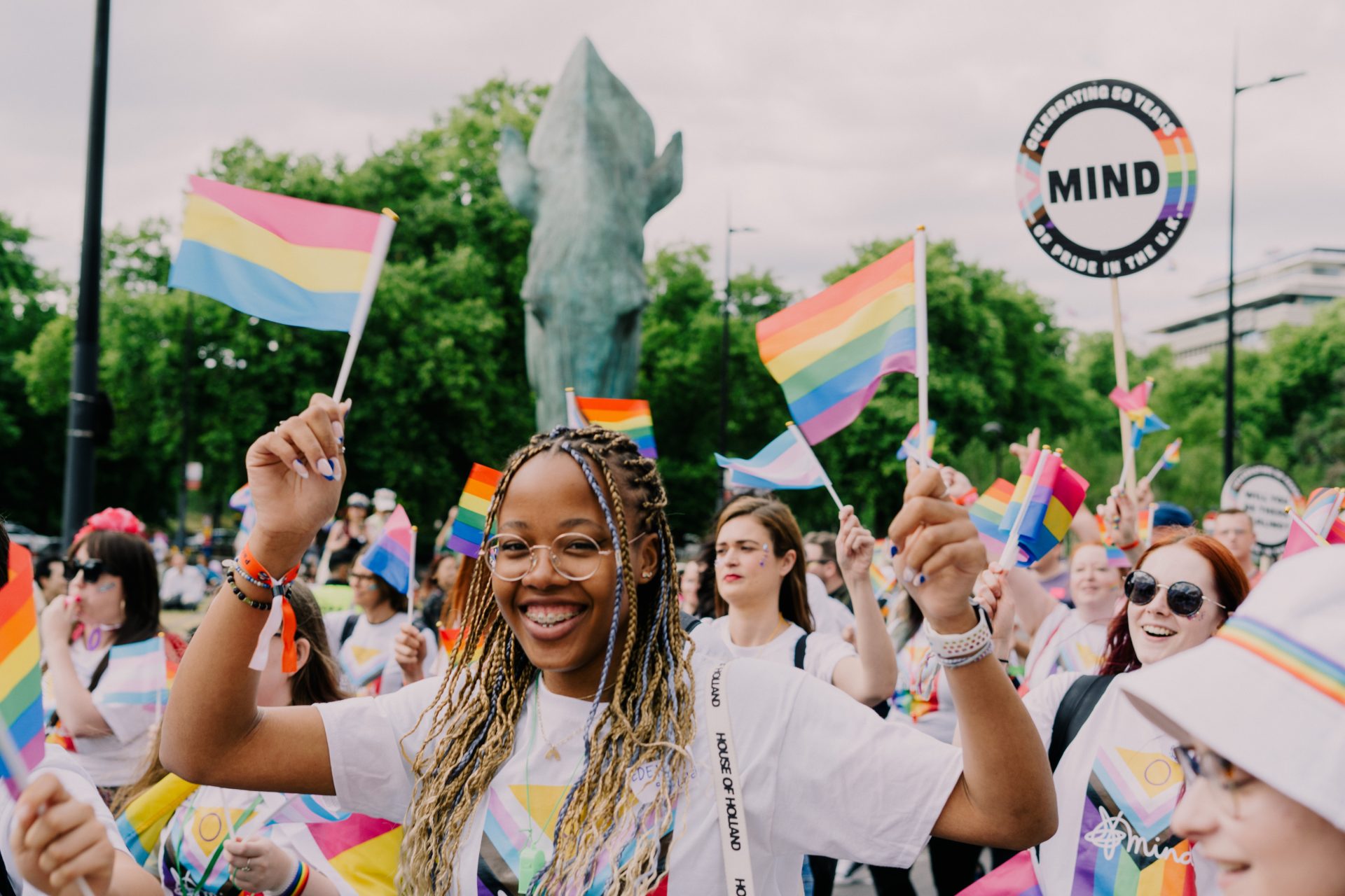 A diverse group of people is seen joyfully participating in a Pride parade. They are waving various Pride flags, including the rainbow flag and the pansexual flag. The individuals are dressed in colorful attire and accessories, with some wearing t-shirts that have Pride-related designs. One prominent figure in the foreground is a smiling young woman with braided hair, glasses, and braces, holding a flag. The atmosphere is festive, with everyone appearing to be in high spirits. In the background, a large statue and lush greenery are visible, suggesting an outdoor setting in a park or public space. Signs, including one from the organisation "MIND," are held high, celebrating Pride.