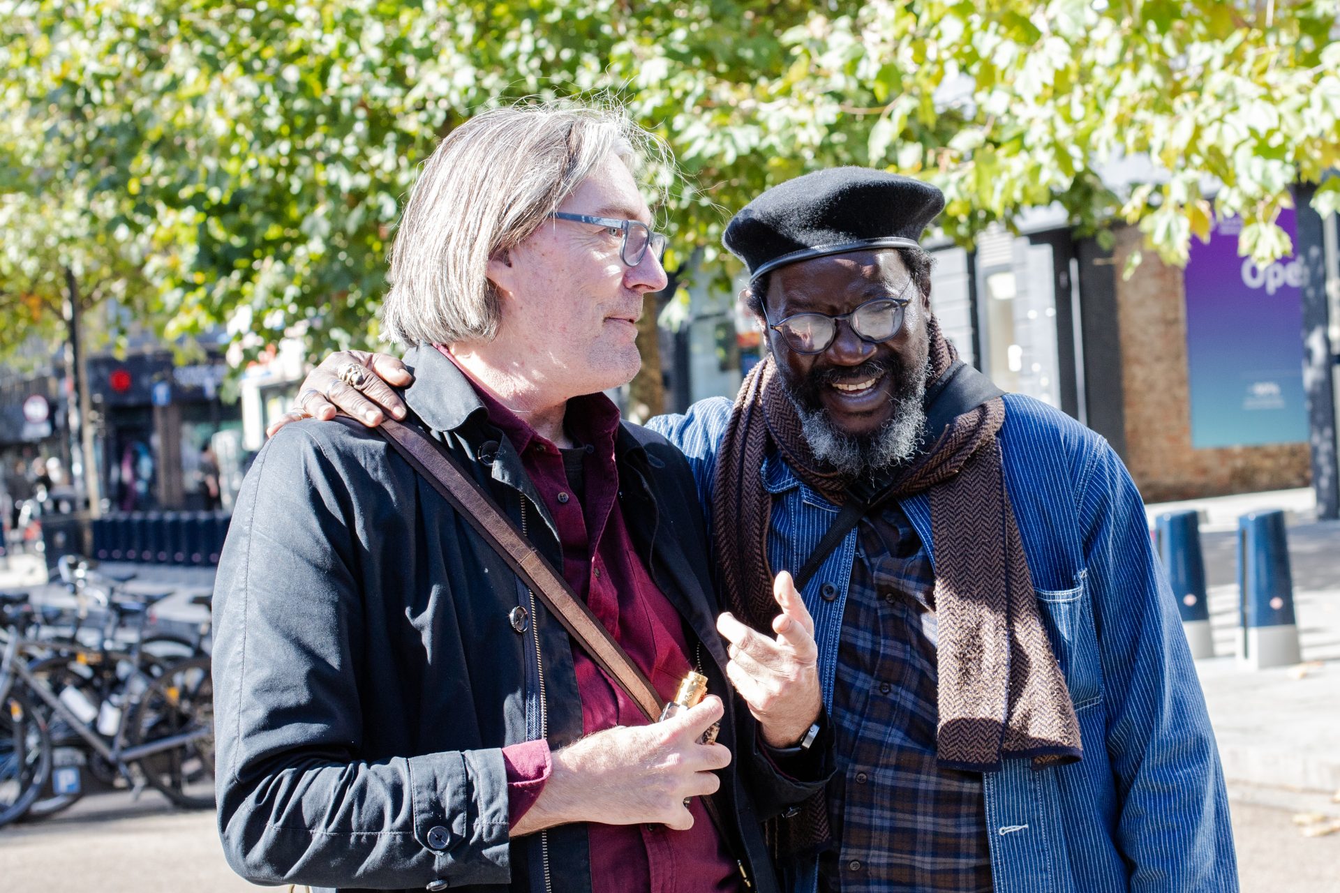 Two men are standing together outdoors in a friendly embrace, with the man on the left wearing glasses, a dark jacket, and a burgundy shirt, holding a small object in his hand. The man on the right, also wearing glasses, has a gray beard and is dressed in a black beret, a blue jacket, and a brown scarf. They are both smiling and seem to be engaged in a pleasant conversation, with a backdrop of trees and an urban setting with bicycles in the background.