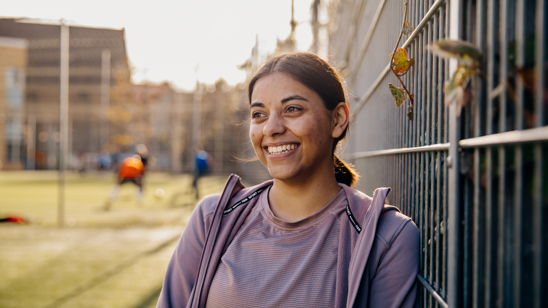 A young woman is leaning against a metal fence, smiling brightly. She is wearing a light purple athletic jacket over a matching top. The background features a sports field with people playing soccer, slightly out of focus, suggesting a sunny day with clear weather. The overall mood of the image is cheerful and relaxed.