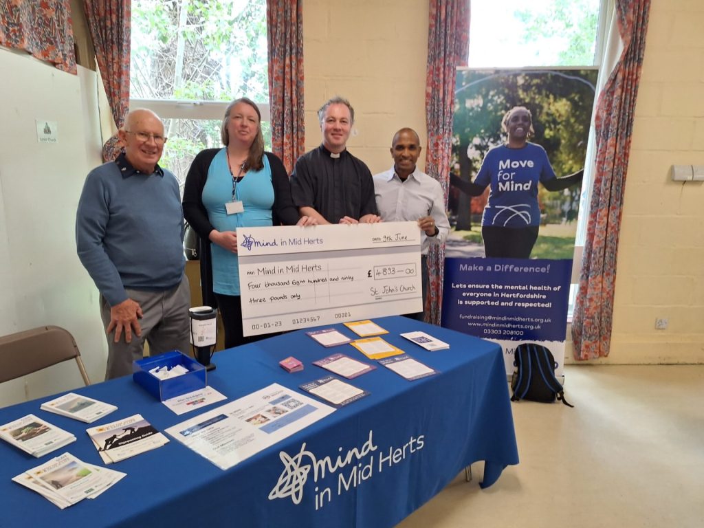 [Image Description: Representatives from St John's Church and Mind in Mid Herts are standing together, holding a large cheque for £4,893. They are smiling in a well-lit room with promotional materials from Mind in Mid Herts on a table in front. The backdrop features a banner promoting mental health awareness and community support.]