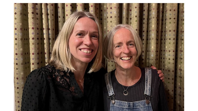 The image shows Caroline and Alison standing side by side, both smiling warmly at the camera. Caroline, on the left, has shoulder-length blonde hair and is wearing a black, textured top. Alison, on the right, has short, light-coloured hair and is wearing denim dungarees over a black shirt. They are in an indoor setting with patterned curtains in the background, radiating a friendly and joyful atmosphere.