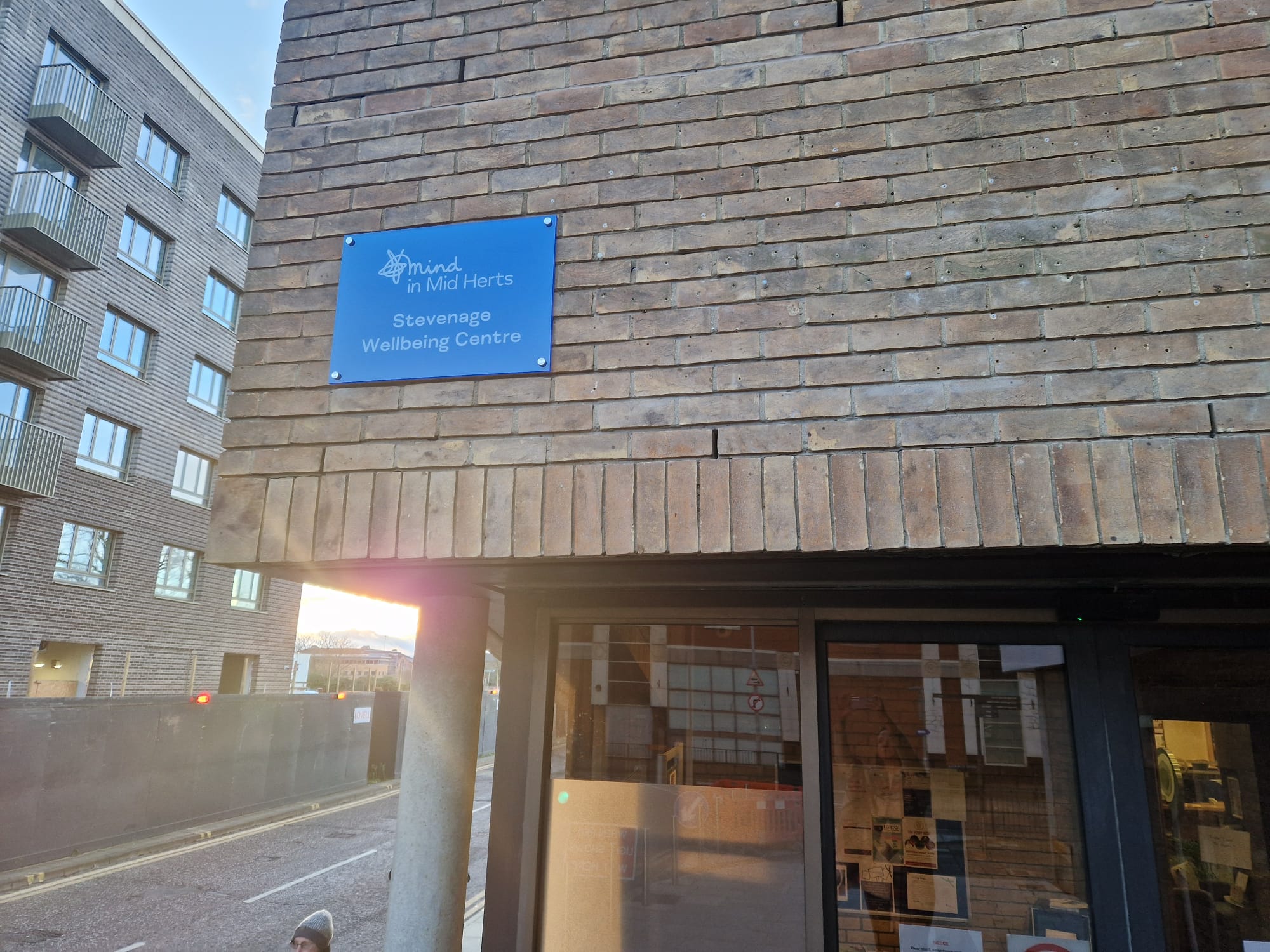 A brick building with a blue sign that reads "Mind in Mid Herts - Stevenage Wellbeing Centre" near the entrance.