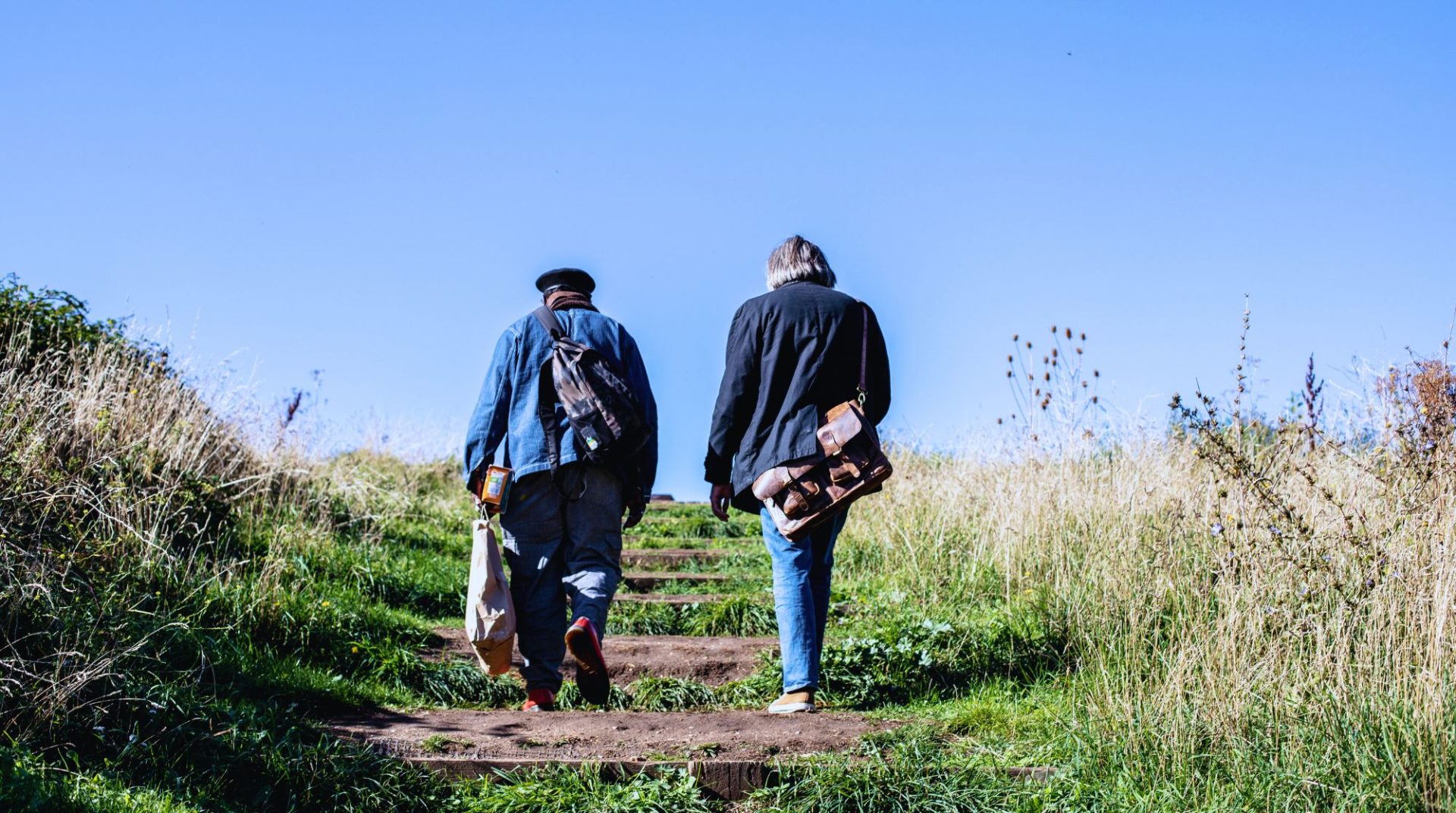 Two people walking up a grassy path surrounded by wild plants under a clear blue sky, each carrying bags and backpacks, symbolising a journey or support.