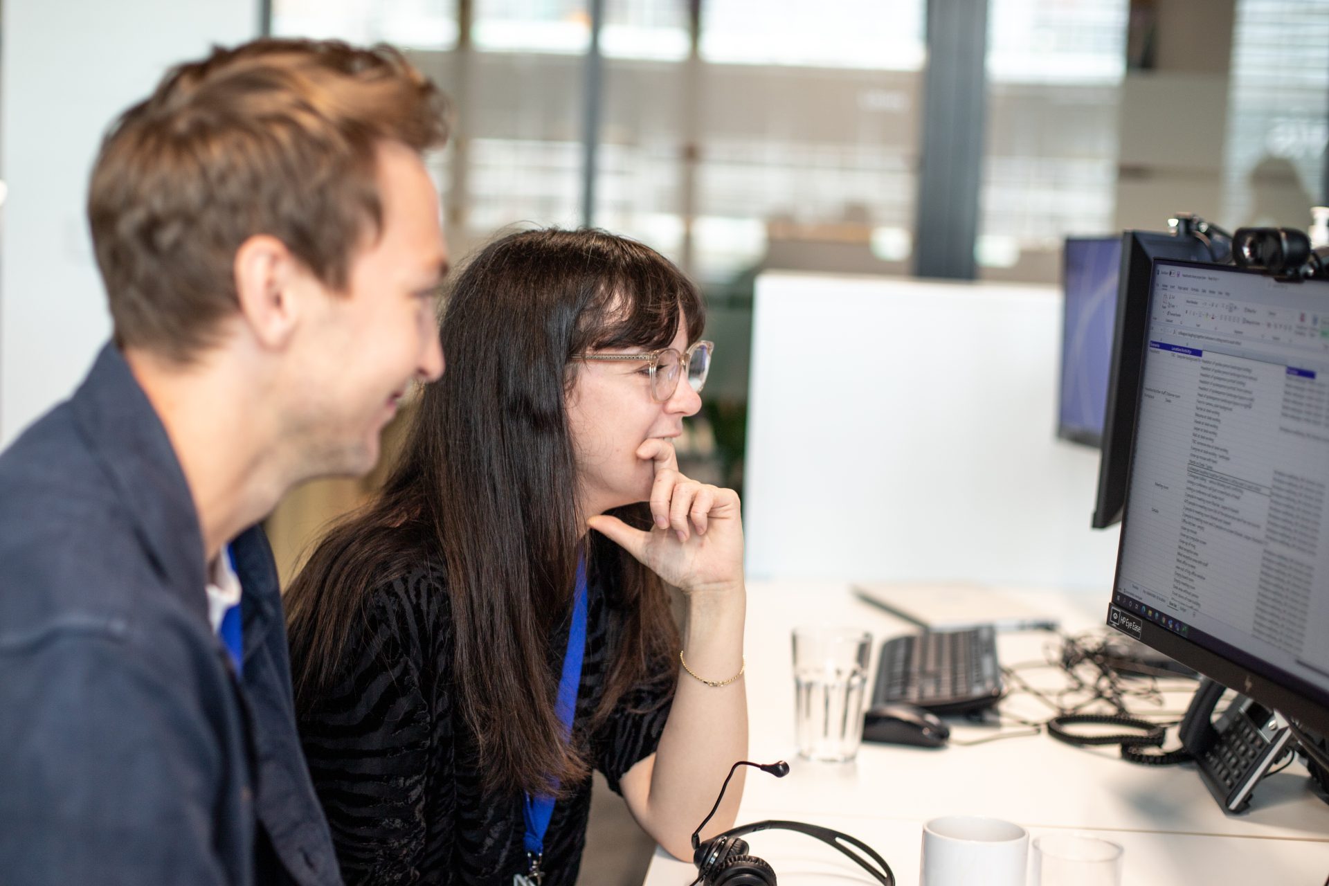 Two colleagues working together at a desk in a modern office, focused on a computer screen displaying a list of data. One person is smiling, while the other is thoughtfully looking at the monitor, wearing glasses and a lanyard. A headset, glass of water, and coffee mugs are visible on the desk.